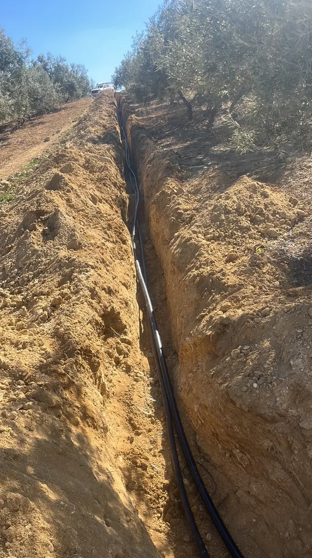 Zanja larga y recta con tuberías negras tendidas, abierta en una ladera de olivos